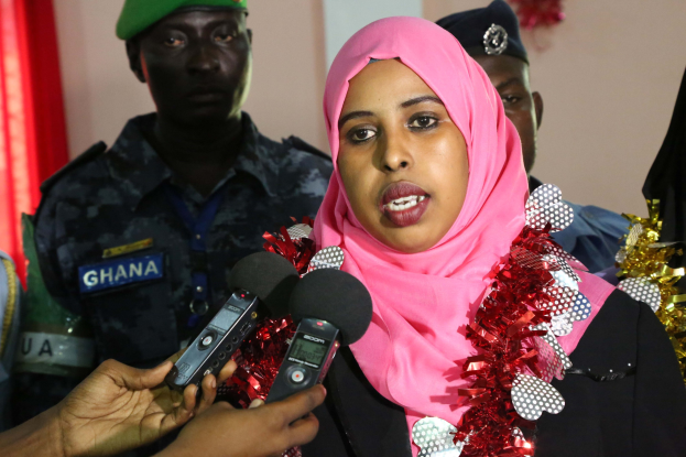 Eine Frau mit einem pinken Kopftuch spricht mit Reportern während eines Interviews, wobei ein Mikrofon links und eine Vorhangwand im Hintergrund zu sehen ist.