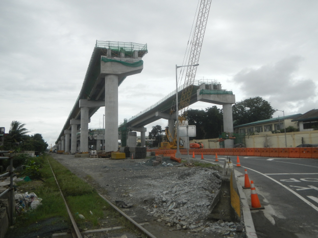 Baustelle mit einer Brücke im Hintergrund, Straße mit Verkehrskegeln markiert, verstreute Steine und Gras, Bahnschiene links, Bäume und Gebäude flankieren die Straße und ein bewölkter Himmel darüber.