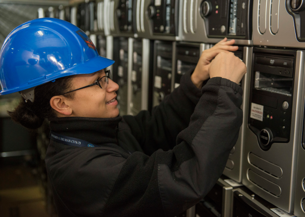Eine Frau in einem blauen Helm und Brille arbeitet an einem Server in einem Rechenzentrum, umgeben von verschiedenen elektronischen Geräten.