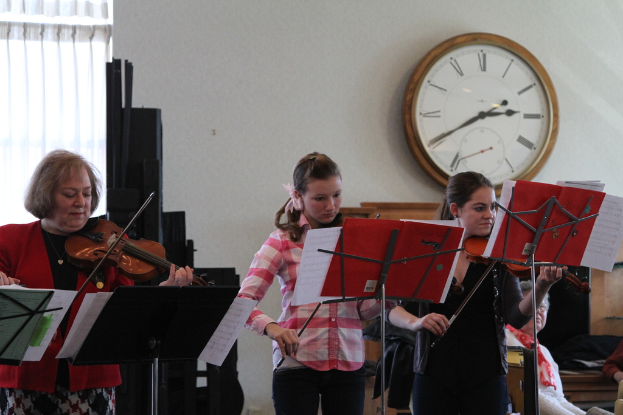 Drei Frauen, die Geigen spielen, auf der Bühne mit Notenpulten vor ihnen, während eine Gruppe sitzender Frauen im Vordergrund zuhört; eine Uhr hängt an der weißen Wand im Hintergrund, mit einem weißen Vorhang in der linken Ecke.