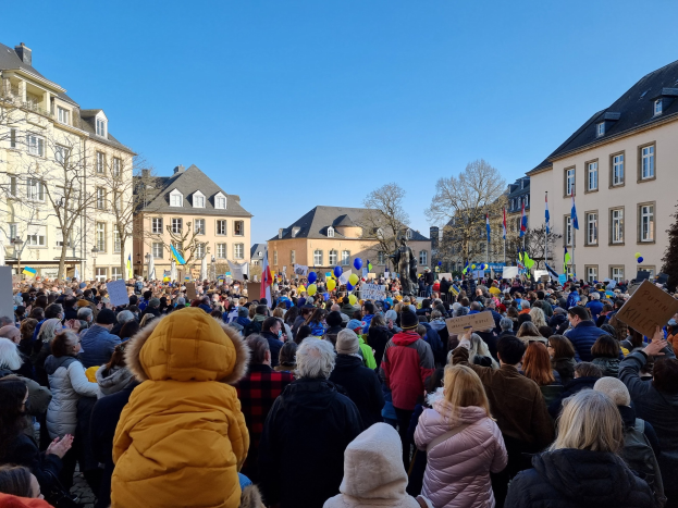 Eine große Menschenmenge protestiert vor einem Gebäude, hält Schilder und Fahnen, mit Bäumen und Fenstern im Hintergrund bei klarem Himmel.