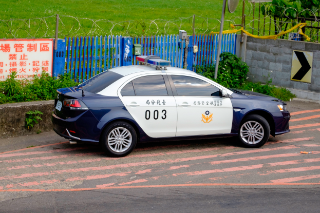 Ein Polizeiwagen am Straßenrand geparkt, umgeben von Pflanzen, einem Zaun, einer Tafel mit Text, einer Wand, einem Pfahl, einer Schüsselantenne und Bäumen.