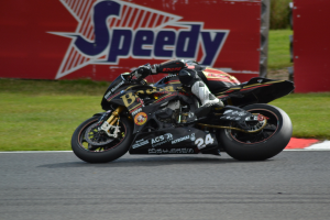 Eine Person in Helm und Schuhen fährt ein Motorrad auf einer Rennstrecke, die von Gras umgeben ist, mit einer Tafel im Hintergrund, auf der Text angezeigt wird.