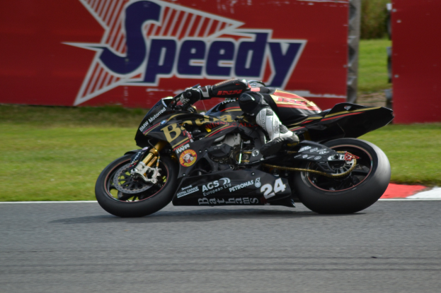 Eine Person in Helm und Schuhen fährt ein Motorrad auf einer Rennstrecke, die von Gras umgeben ist, mit einer Tafel im Hintergrund, auf der Text angezeigt wird.