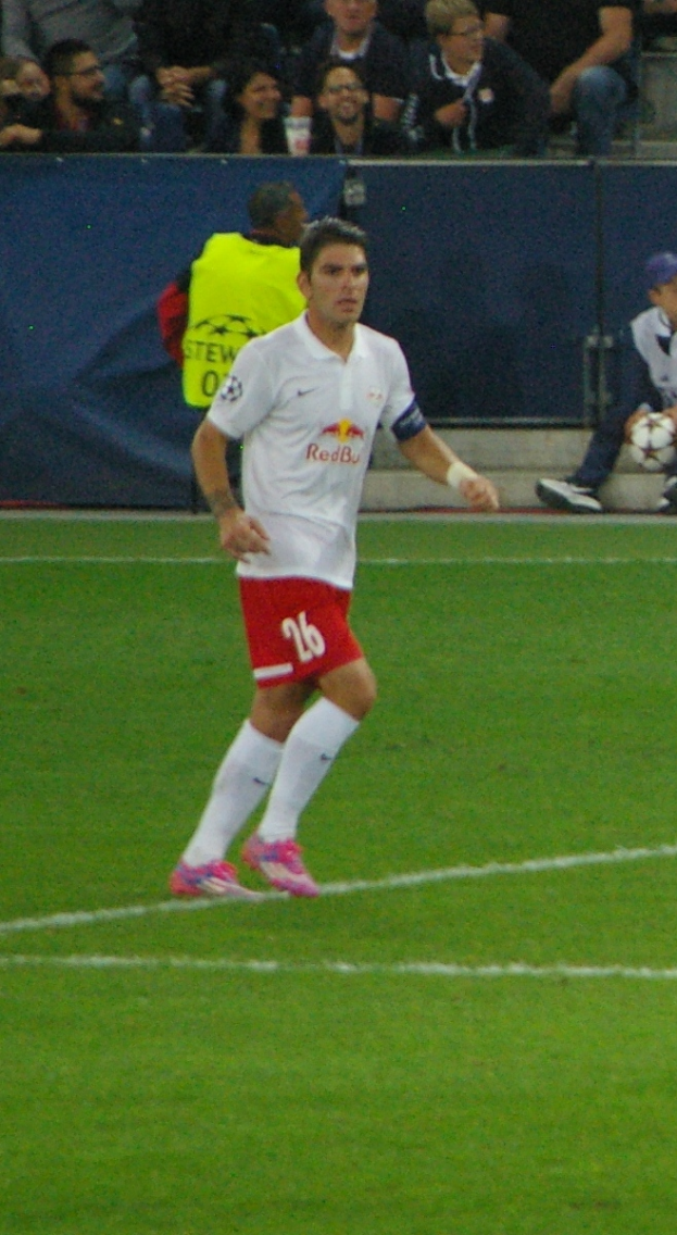 Ein Mann in einem weißen Hemd und roten Shorts spielt Fußball auf einem Feld, umgeben von einer Gruppe von Menschen, die im Hintergrund sitzen.