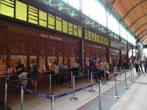 Eine Gruppe von Menschen steht in einer Warteschlange an einem Bahnhof, einige tragen Taschen, mit Stangen mit Bändern, Schildern, Deckenlampen und Treppen mit Geländern und einem Gebäude im Hintergrund.