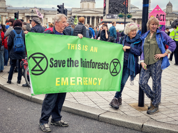 Eine Gruppe von Menschen, die auf einem Fußweg stehen und ein grünes Banner halten, auf dem 'Regenwaldrettung Notfall' steht, mit Gebäuden, Pfählen, Verkehrszeichen und einem klaren blauen Himmel im Hintergrund.