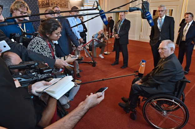 Ein Mann im Rollstuhl umgeben von Reportern mit Kameras, Mikrofonen und Handys sowie einem Banner, Lichtern und einer Wand im Hintergrund.