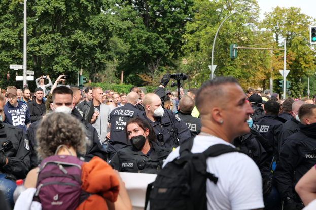 Eine große Gruppe von Menschen steht vor einer Reihe von Polizei Beamten, einige tragen Masken und haben Rucksäcke oder Kameras dabei, mit Laternenmästen, Verkehrsampeln, Schildern, Bäumen und einem klaren blauen Himmel im Hintergrund.