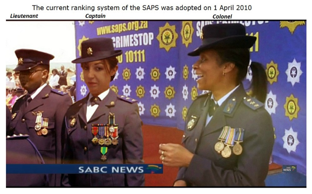 Gruppe von Menschen in Uniformen und Hüten mit einem Banner, Text oben und unten mit der Aufschrift "Das aktuelle Rangsystem der SAP wurde am 1. April 2010 übernommen."