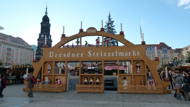 Ein lebendiger Weihnachtsmarkt in Dresden, Deutschland, mit Menschen, die gehen, Taschen tragen und ein Schild mit Text, vor dem Hintergrund von Bäumen, Gebäuden und einem bewölkten Himmel.