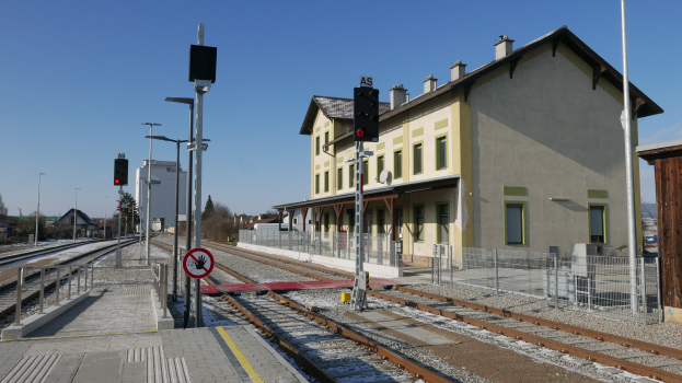 Ein Bahnhof mit einem Zug auf den Schienen, umgeben von Gebäuden, Straßenlaternen, Verkehrspfosten, Verkehrssignalen, Schildern, Zäunen, Bäumen und einem klaren blauen Himmel.