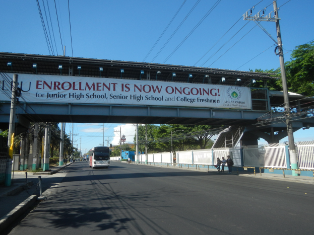 Ein Bus fährt unter einer Brücke mit einem Schild, auf dem "Jetzt Anmeldung" steht, vorbei, mit Menschen auf dem Gehweg darunter, Strommasten mit Drähten an der Straße und Bäumen mit einem klaren blauen Himmel im Hintergrund.