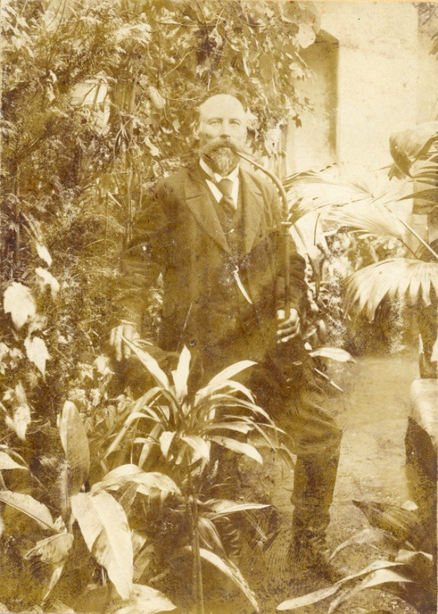 Altes Schwarz-Weiß-Foto eines Mannes in Anzug und Krawatte, der in einem Garten mit Pflanzen und Bäumen steht, mit einer Wand im Hintergrund.