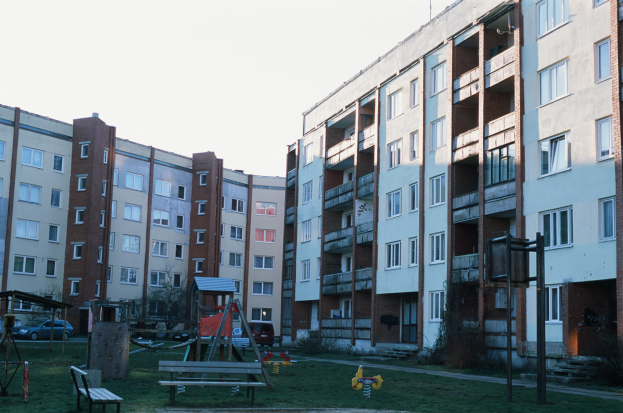 Wohngebäude mit Spielplatz, Bänken, Schaukeln, Gras, Bäumen, Fahrzeugen und einem klaren blauen Himmel im Hintergrund.
