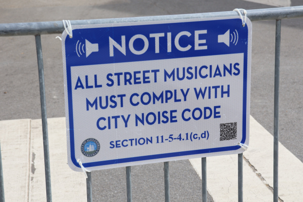 Anschlagtafel an einem Zaun mit einem Schild, auf dem "Anschlag: Alle Straßenmusiker müssen sich an die Stadtlärmvorschriften halten" steht, und eine Straße im Hintergrund.