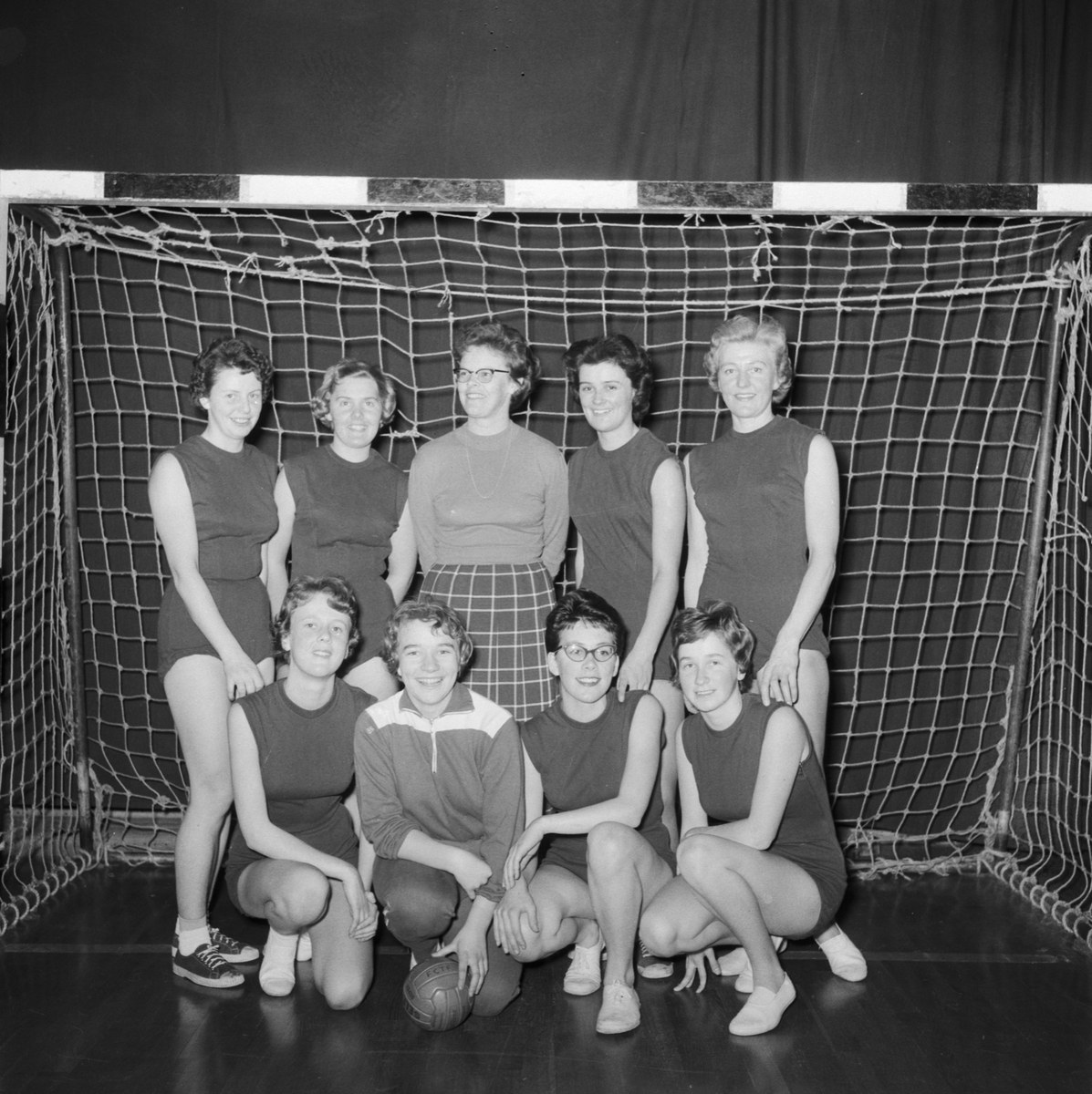 Schwarzes und weißes Foto einer Gruppe von Frauen, die vor einem Fußballtor posieren, mit einem Ball auf dem Boden und einem Vorhang im Hintergrund.