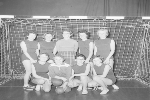 Schwarzes und weißes Foto einer Gruppe von Frauen, die vor einem Fußballtor posieren, mit einem Ball auf dem Boden und einem Vorhang im Hintergrund.