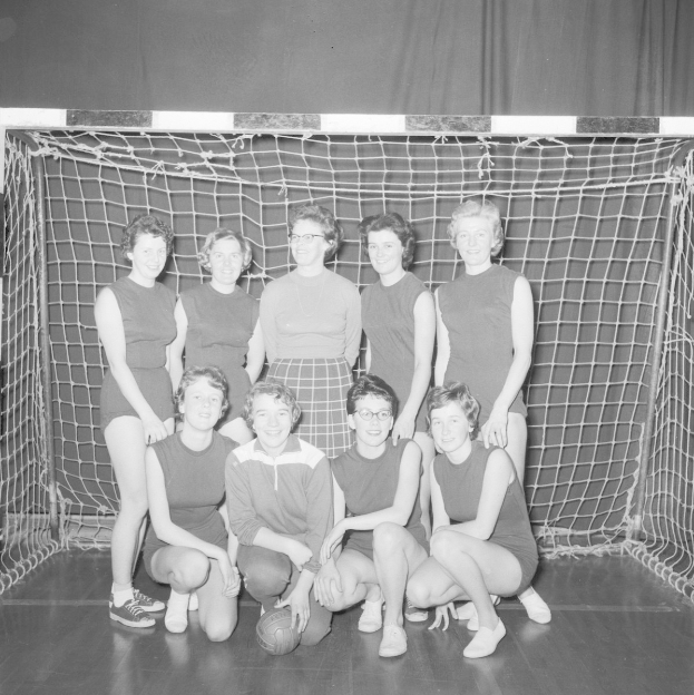 Schwarzes und weißes Foto einer Gruppe von Frauen, die vor einem Fußballtor posieren, mit einem Ball auf dem Boden und einem Vorhang im Hintergrund.