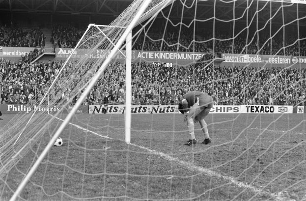 Ein Schwarz-Weiß-Foto eines Fußballspielers, der vor einer Zuschauermenge im Stadion einen Ball hält, mit einem Torpfosten und einem Ball in der Nähe und Schilder mit Text im Hintergrund.