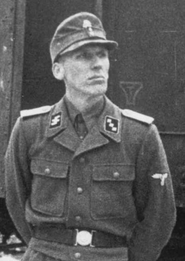 Ein Schwarz-Weiß-Foto eines Mannes in Uniform, der eine Mütze trägt und vor einem Zug steht, mit Text am unteren Rand, der ihn als deutschen Soldaten ausweist.