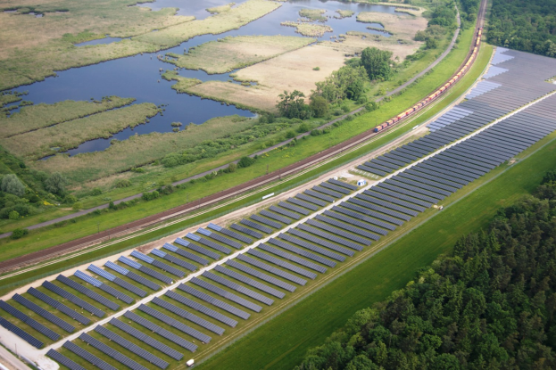 Luftaufnahme eines Solarparks in einem Feld mit Solarpanelen, umgeben von Bäumen, Gras, Pflanzen und Wasser, mit einem Zug auf einer näheren Eisenbahnschiene.