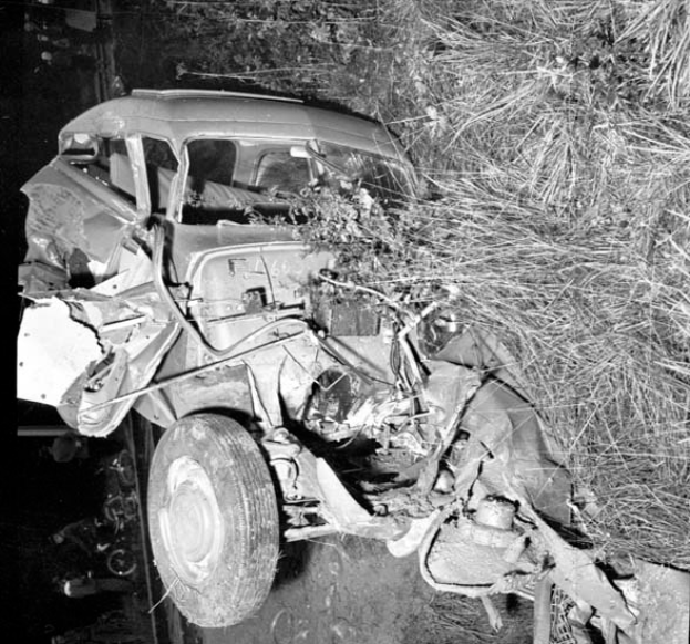 Ein Schwarz-Weiß-Foto eines schwer beschädigten Autos am Straßenrand mit einer eingedrückten Front, zersplitterten Fenstern, einem Baum im Hintergrund und verstreuten Gegenständen auf der Szene.