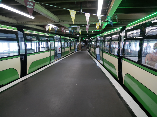Grüner und weißer Zug fährt auf Schienen neben einem Bahnsteig mit sichtbaren Passagieren, unter einer Decke mit Lichtern und dekorativen Flaggen an einem Münchner Bahnhof.