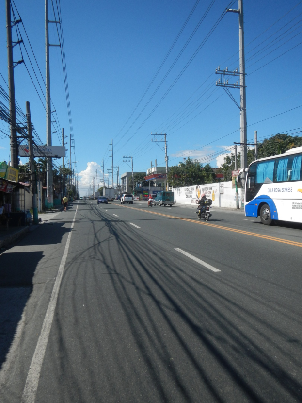 Ein Bus fährt auf einer Straße mit hohen Strommasten und Drähten, flankiert von Geschäften und Fußgängern auf der linken Seite, mit Gebäuden, Bäumen, einer Wand und einem bewölkten Himmel im Hintergrund.