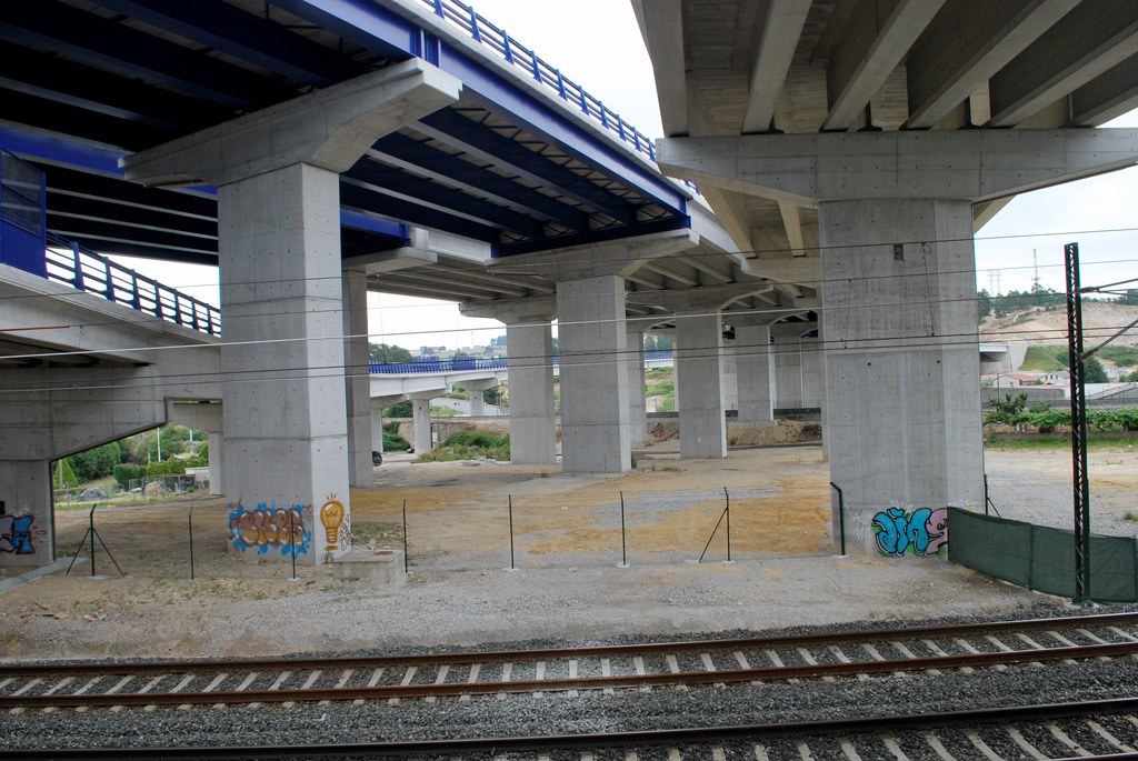 Eine Bahnschiene mit Graffiti an der Seite, die unter einer von Pfeilern und Geländern gestützten Brücke hindurchführt, mit Strommasten und Oberleitungen daneben, vor einem Hintergrund aus Bäumen, Gebäuden und einem klaren blauen Himmel.