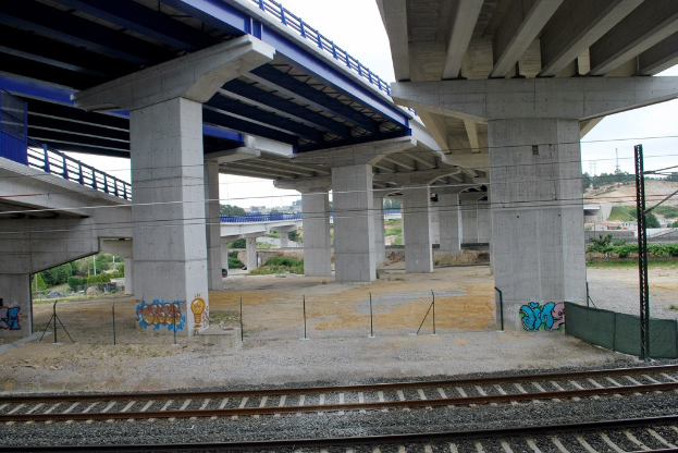 Eine Bahnschiene mit Graffiti an der Seite, die unter einer von Pfeilern und Geländern gestützten Brücke hindurchführt, mit Strommasten und Oberleitungen daneben, vor einem Hintergrund aus Bäumen, Gebäuden und einem klaren blauen Himmel.