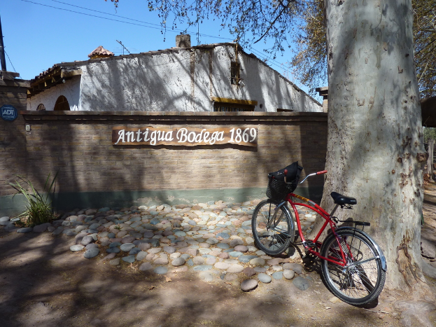Ein rotes Fahrrad steht auf einem großen Baumstamm mit Steinen auf dem Boden, mit einer Begrenzungsmauer, auf der "Antike Bodega 1869" steht, und einem weißen Haus im Hintergrund.