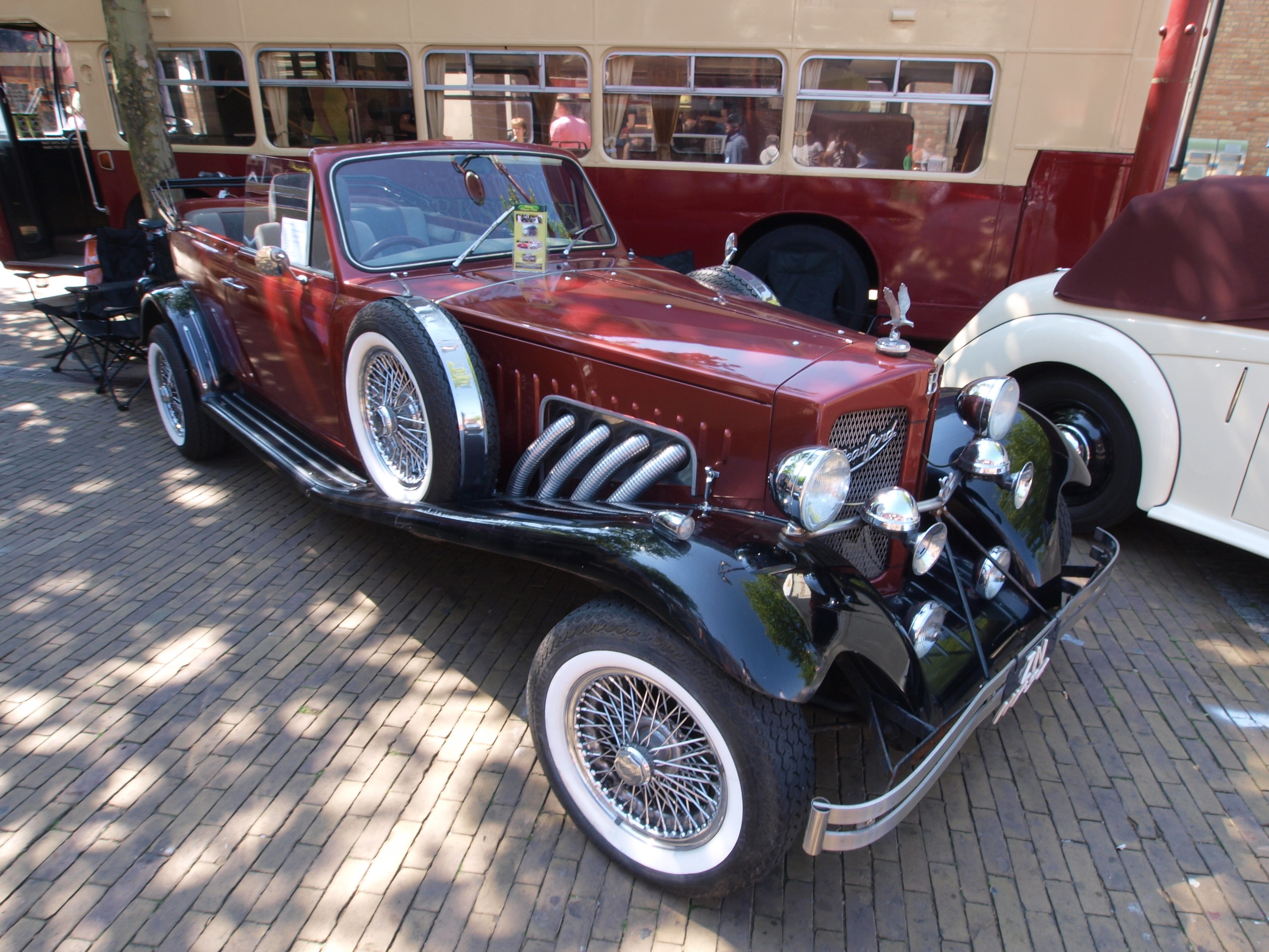 Ein Oldtimer neben einem Doppeldeckerbus auf Kopfsteinpflasterstraße, mit Stühlen und einem Baum im Vordergrund und einem Gebäude im Hintergrund.