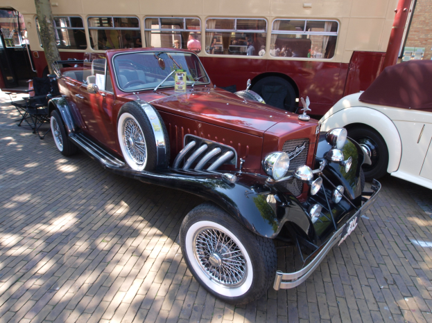 Ein Oldtimer neben einem Doppeldeckerbus auf Kopfsteinpflasterstraße, mit Stühlen und einem Baum im Vordergrund und einem Gebäude im Hintergrund.