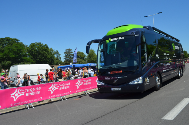 Ein schwarzer und grüner Bus fährt auf einer Straße neben einer Menschenmenge, einige tragen Mützen, mit einem Banner auf der linken Seite und Bäumen und einem klaren blauen Himmel im Hintergrund.