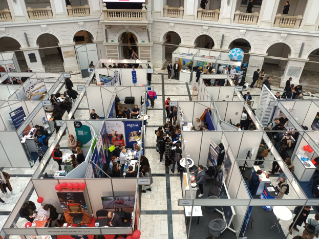 Große Gruppe von Besuchern auf einer internationalen Fachmesse mit Ständen, Bannern, Tischen, Stühlen und Luftballons in einem Raum mit architektonischen Bögen, Säulen und einem Bildschirm im Hintergrund.