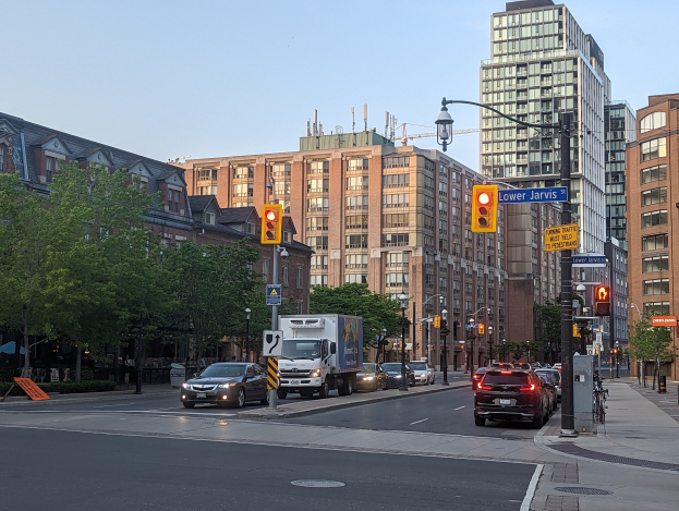 Eine städtische Straße mit starkem Verkehr einschließlich eines Lastwagens, eines Autos und eines SUV, gesäumt von Laternenmasten, Verkehrsampeln, Texttafeln, Bäumen und Gebäuden unter einem Himmel.
