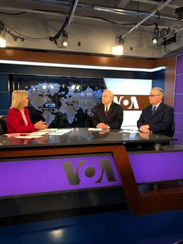 Drei Personen, zwei Männer in Blazern und Krawatten und eine Frau in einem roten Kleid, sitzen an einem Schreibtisch mit Papieren und Gegenständen, diskutieren vor einem Fernseher, mit einem Bildschirm und Lichtern im Hintergrund.