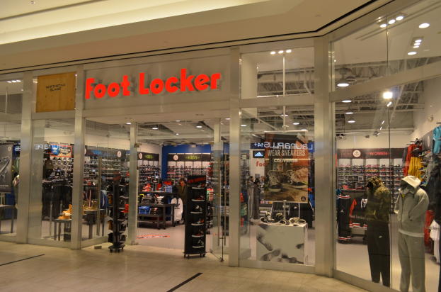Außenansicht eines Foot Locker Ladens in einem Einkaufszentrum mit Glaswänden und -türen, Schaufensterpuppen in Kleidern, Regalen mit Schuhen und Kleidung sowie Deckenbeleuchtung.