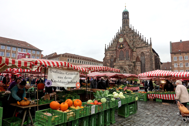 Ein belebter Markt in Nürnberg, Deutschland, mit Ständen, die verschiedene Früchte und Gemüse anbieten, Menschen mit Taschen und aufgestellten Zelten, sowie Gebäuden und einem Uhrenturm im Hintergrund unter einem sichtbaren Himmel.