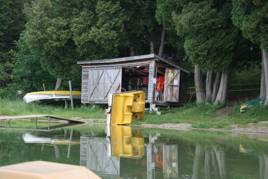 Ein Boot auf dem Wasser neben einem Schuppen mit einigen Gegenständen, umgeben von Bäumen, einem Zaun und anderen Objekten.