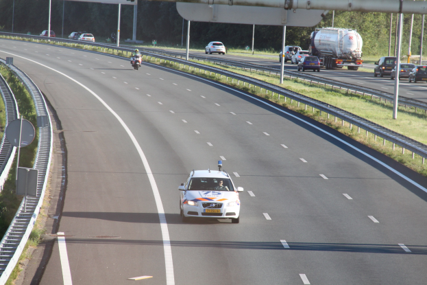 Ein Auto auf einer Autobahn mit einem stationären Polizeiwagen auf dem Seitenstreifen, andere Fahrzeuge und Bäume, Geländer, Pfosten und Bretter im Hintergrund.