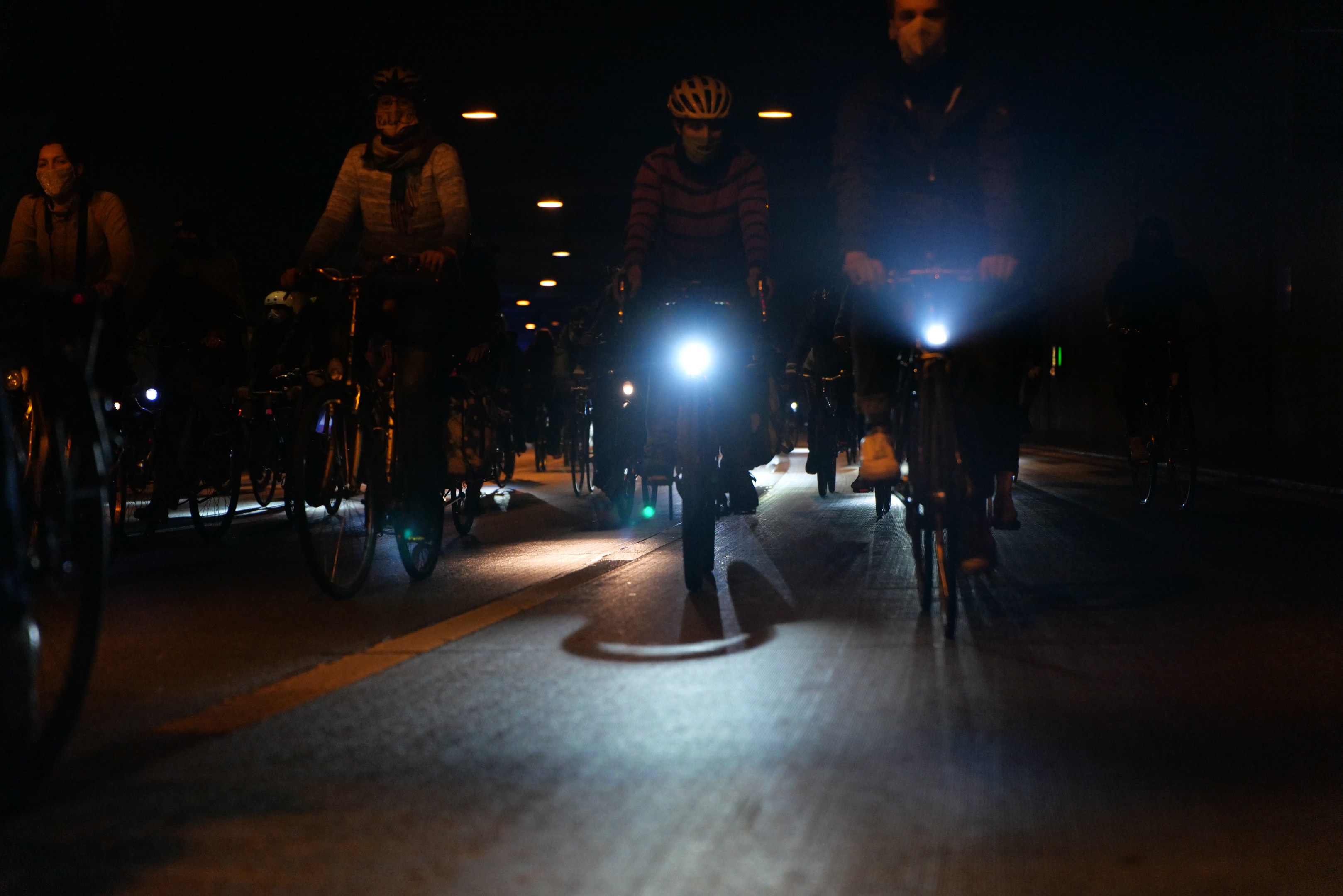 Gruppe von Menschen in Helmen auf Fahrrädern auf einer nächtlichen Stadtstraße unterwegs, beleuchtet von urbanen Lichtern.