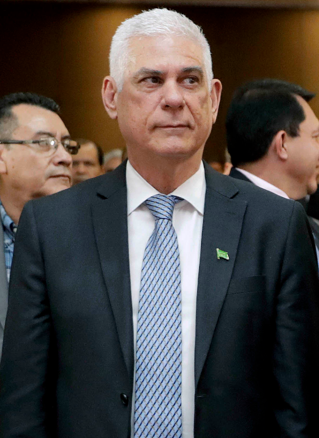 Ein Mann in einem marineblauen Anzug, weißem Hemd und gemustertem Schlips steht vor einer Gruppe von Menschen und sieht ernst aus, mit ordentlich gekämmtem Haar und einer Wand im Hintergrund.