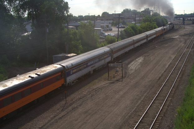 Ein Zug auf Schienen mit Bäumen, Strommasten mit Kabeln, Autos und Gebäuden zur Linken und einer Brücke vorne, die Rauch abgibt.