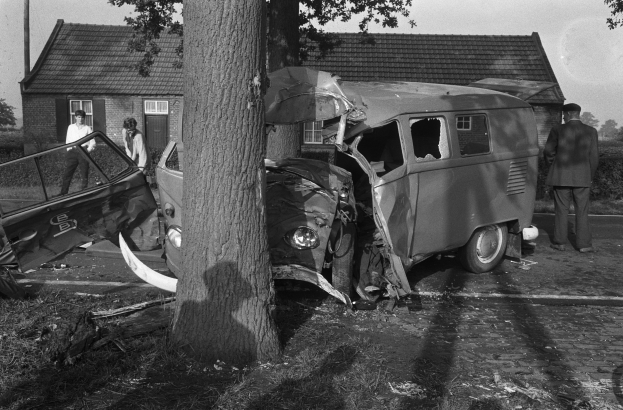 Schwarz-weißes Bild eines verunglückten Autos am Straßenrand mit einem Baum im Vordergrund, einigen Menschen in der Nähe und Häusern, Bäumen und einem Pfahl im Hintergrund.