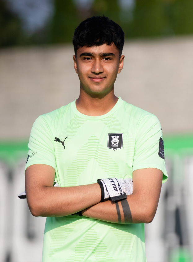 Ein junger Mann in einem grünen Hemd steht auf einem Fußballfeld und verschränkt die Arme, während er lächelt, mit einem unscharfen Hintergrund.