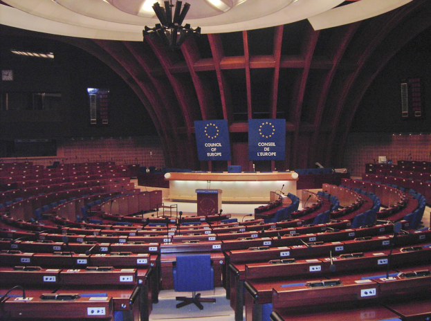 Der Sitzungssaal des Europäischen Parlaments in Brüssel mit einer halbrunden Sitzanordnung, einem zentralen Podium, an der Wand montierten Texttafeln und einer beleuchteten Decke.