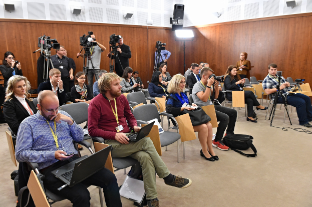 Eine Gruppe von Menschen, die mit Laptops, Kameras und Taschen in einem Konferenzraum mit Lampen und Lautsprechern im Hintergrund sitzen.