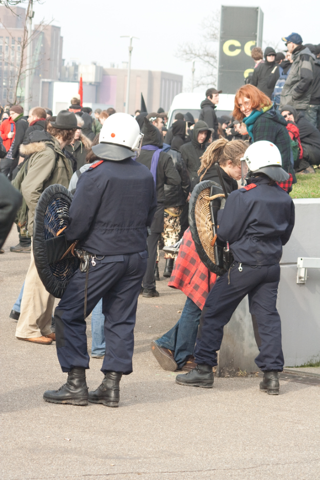 Eine Gruppe von Menschen, die auf der Straße gehen, mit zwei Personen vorne, die wie Polizisten aussehen, Gebäuden im Hintergrund und Boden unten.
